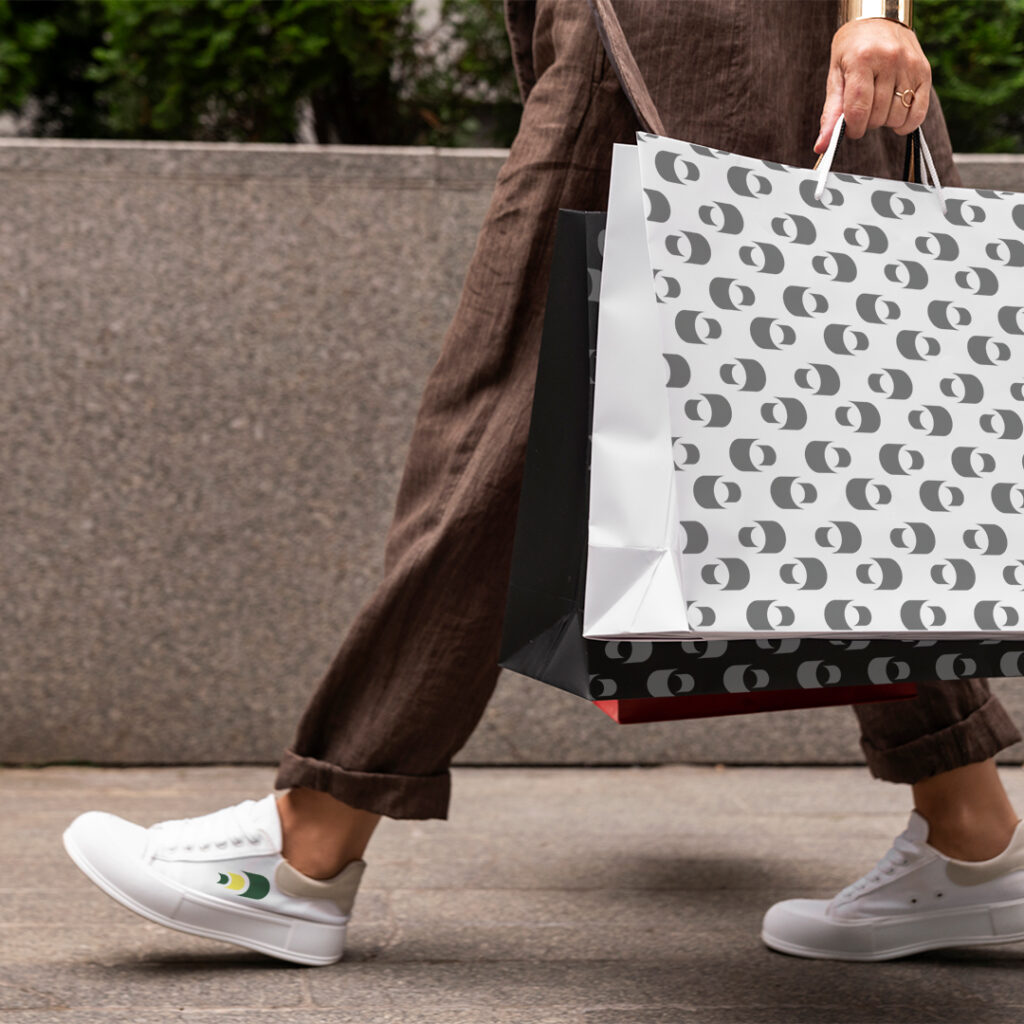 Mockup di un borsa da shopping in carta per il brand Centipede, trasportata a mano da una persona che indossa pantaloni marroncini, scarpe bianche con marchio Centipede, su un marciapede in cemento